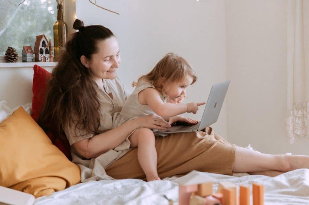 Side view of woman with curious small girl in dress looking at laptop while sitting together in bedroom